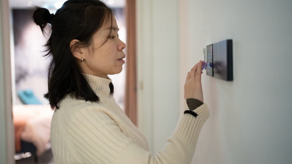 A young woman wearing a white sweater adjusts a smart thermostat on the wall of her home